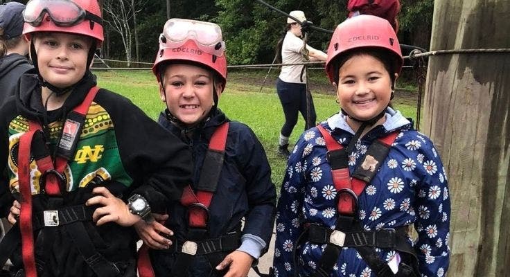 Three students at camp wearing safety helmets and harnesses getting ready for an activity