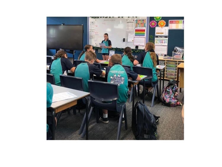 students in a classroom sitting at desks while one student stands up the front speaking
